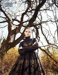 a woman standing in front of a tree wearing a black dress with gold embroidery on it