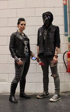 two people standing next to each other in front of a white brick wall with gas pumps