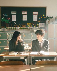 two young people sitting at desks in a classroom with books and papers on them