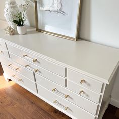 a white dresser with gold handles and knobs on the drawers, in front of a framed art piece