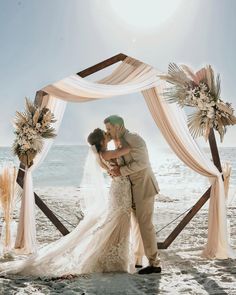 a bride and groom kissing under an arch on the beach