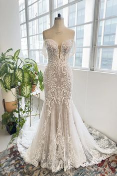 a wedding dress on display in front of a large window with potted plants behind it