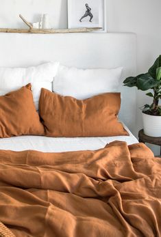 a bed with orange sheets and pillows in a white room next to a potted plant