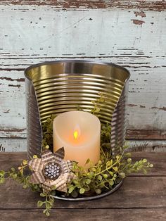 a lit candle in a metal container filled with greenery and leaves on a wooden table