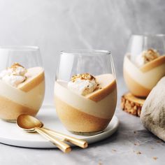 two glasses filled with dessert sitting on top of a white plate next to gold spoons