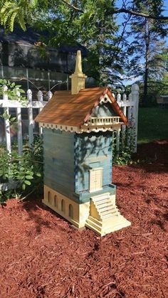 a bee house sitting in the dirt next to a white picket fence and some trees