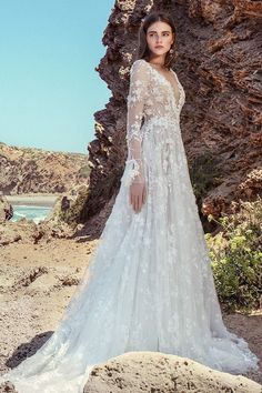 a woman in a white wedding dress standing on rocks near the ocean with her arms behind her back