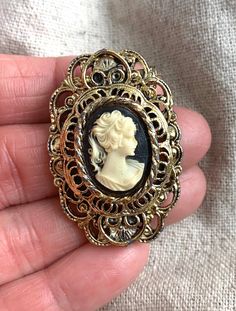 "You will receive this vintage cameo brooch from Gerry's. It measures 1 3/4\" x 1 1/8\" and is cast in an antiqued gold tone base metal. The cameo backing is black and the silhouette is a cream color. This brooch is in very good vintage condition with minimal signs of wear and a properly functioning safety catch. For finished vintage assemblage and vintage inspired necklaces, bracelets, and earrings, please visit us at our other Etsy store, William Dalton Design: www.williamdaltondesign.etsy.com Vintage Medallion Brooch For Wedding, Vintage Medallion Brooches For Wedding, Antique Gold Medallion Brooch, Antique Gold Brooches For Vintage Collection, Antique Gold Brooches From Vintage Collection, Antique Gold Brooch Jewelry, Ornate Medallion Brooches For Collectibles, Gold Antique Brooches For Vintage Collection, Ornate Medallion Brooches For Collectors