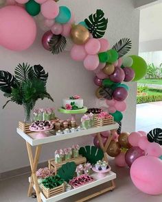 a table topped with lots of balloons and desserts next to a wall filled with palm leaves