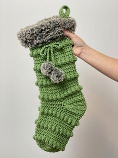 a hand holding up a green crocheted stocking with grey pom - poms