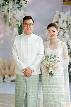 a man and woman standing next to each other in front of a floral wall with white flowers