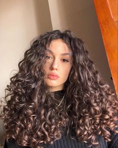 a woman with long curly hair standing in front of a wooden door wearing a black sweater