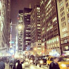 many people are crossing the street at night in new york city, with skyscrapers lit up