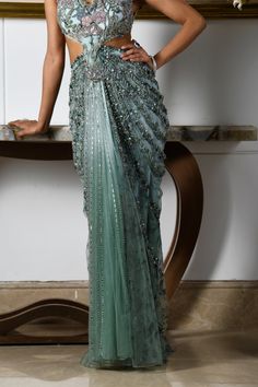 An eye-catching ensemble immersed in the marine allure of aqua blue creates an elegant look. This modern concept saree features a sleeveless blouse and a pre-draped skirt attached to each other. Botanical motifs, embroidered with fine crystals, sequins, and beads, adorn the entire ensemble, along with tulle pleats. The beauty of the wilderness perfectly interplays its role through flower motifs and deep hues, making it an exquisite piece for your nightly soirees. The sleeveless blouse features signature flower motifs embroidered with the use of crystals, beads, and sequins. Pre-draped skirt adorned with flower motifs attached to the blouse. Criss-cross pattern at the backside of the blouse. The blouse contains a metal zip at the backside. The skirt is attached to the blouse. From Moledro’s Concept Saree, Saree Skirt, Silk Drapes, Botanical Motifs, Lehenga Designs Simple, Crystals Beads, Flower Motifs, Draped Skirt, The Marine