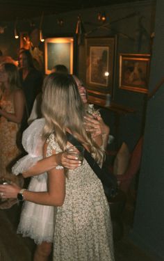 two women hugging each other in a room full of people at a party with paintings on the walls