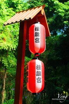 two red and white lanterns hanging from a wooden structure in front of some green trees