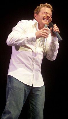 a man holding a microphone in his right hand and smiling at the camera while standing on stage