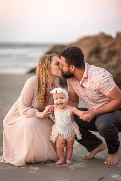 a man and woman kiss their baby on the beach while she kisses her face in front of them