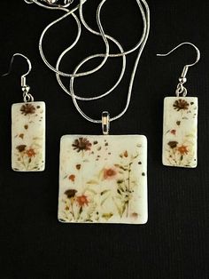 three pieces of jewelry with flowers on them sitting on a black surface, one piece has a silver chain and the other part has a white square shaped pendant