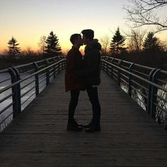 two people standing on a bridge kissing each other with the sun setting in the background