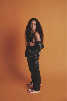 a woman with long hair standing in front of an orange wall wearing black leather pants