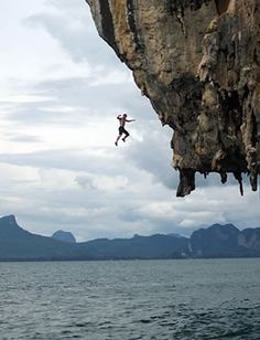 a man jumping off the side of a cliff into the ocean with mountains in the background