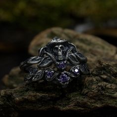 a skull ring sitting on top of a rock next to some leaves and rocks with purple stones