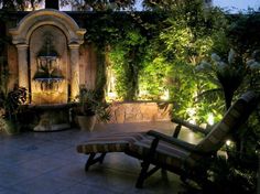 an outdoor patio area with benches and lights on the wall, surrounded by greenery