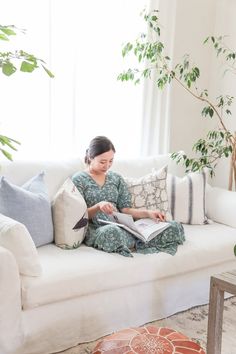 a woman sitting on a couch reading a book in front of a window with potted plants