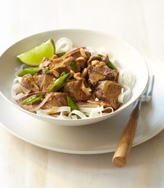 a white bowl filled with noodles and meat on top of a table next to a wooden spoon