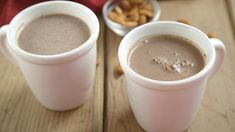 two white cups filled with hot chocolate on top of a wooden table next to nuts