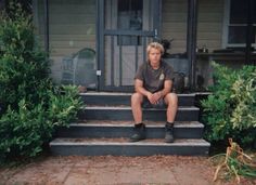 a young man sitting on steps in front of a house with bushes and shrubs around him