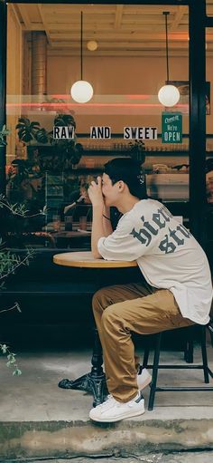 a young man sitting at a table in front of a store window with his hand on his face