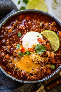 a bowl filled with chili and cheese topped with sour cream, lime wedges and cilantro