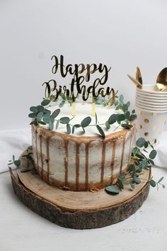 a birthday cake sitting on top of a wooden slice with greenery around it and the words happy birthday written in gold