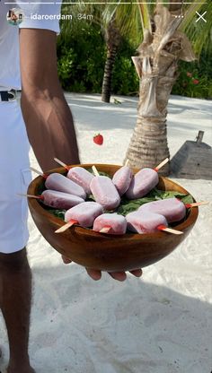 a man holding a wooden bowl filled with food on top of a sandy beach next to palm trees