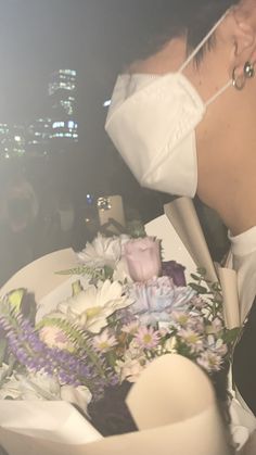 a woman wearing a face mask and holding a bouquet in front of her face with the city lights behind her