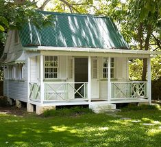 a small white house with a green roof and porch on the grass in front of some trees