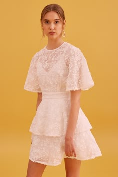 a woman standing in front of a yellow background wearing a white dress with ruffles