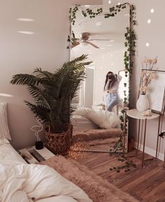 a woman standing in front of a mirror next to a bed and plant on the floor