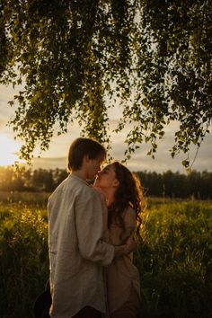 a man and woman kissing under a tree in the middle of a field at sunset