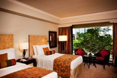 two beds in a hotel room with red and orange pillows on the headboards, one is facing a large window