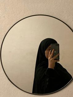 a woman wearing a hijab is looking at her cell phone in the mirror