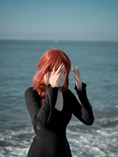 a woman with red hair covers her eyes from the sun by the ocean's edge