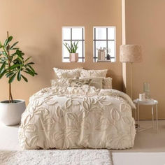 a white bed sitting next to a tall plant in a room with two windows and a rug on the floor