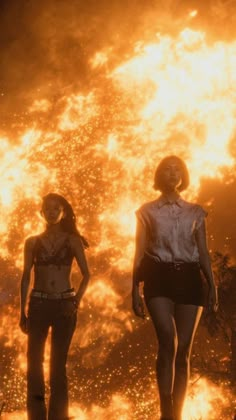 two women standing next to each other in front of a large fire with flames behind them