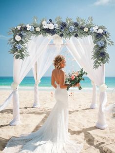 a woman standing on top of a sandy beach under a white wedding arch covered in flowers