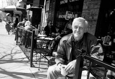 an old man sitting on a bench in front of a building with tables and chairs