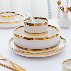 white and gold dinnerware set on a table with flowers in vases behind it