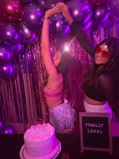 two women in bikinis standing next to a cake with purple and silver balloons behind them